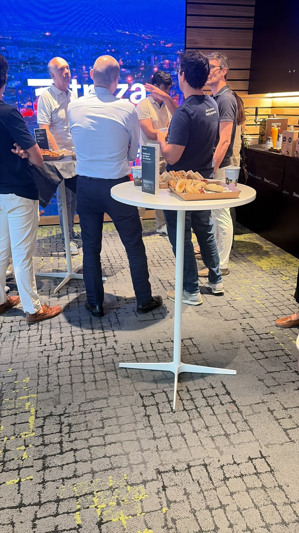 Asistentes conversando de pie junto a una mesa alta, con el logo de Traza en la pantalla del fondo.